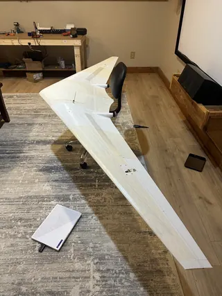 White 3D-printed swept flying wing resting on an office chair indoors, spanning the room, with a workbench in the background