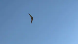White swept flying wing seen from below against a clear blue sky, banking, with its two rear propellers visible