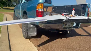 Handheld clip of a white flying wing laid across a pickup tailgate with its hatch off, as the split elevon surfaces on the trailing edge move up and down