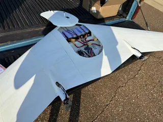 Flying wing on a pickup tailgate with the top hatch lifted, exposing batteries, wiring and electronics; two rear motors visible
