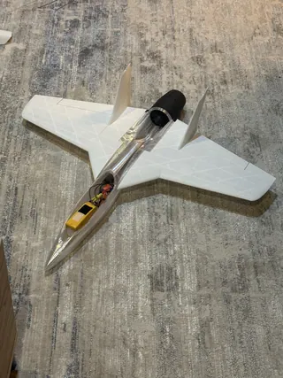 White 3D-printed jet with twin canted fins and a clear fuselage showing wiring and a yellow battery, black ducted fan at the tail