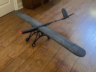 Dark grey 3D-printed high-wing plane with a long rounded-tip wing, slim tail boom, red propeller and wheeled landing-gear frame