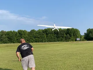 White twin-boom UAV with a long straight wing climbing over a grass field as a person follows through from a hand launch
