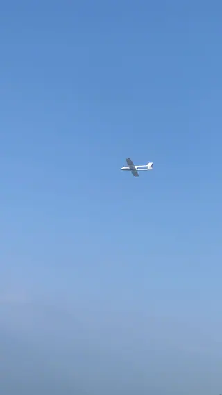 White twin-boom UAV with a long wing banking over a tree line at a flying field